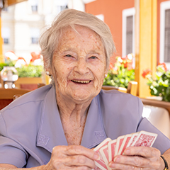 Natural Pharmaceuticals Hepactum FORTE Idős nő mosolyogva kártyázik napsütéses teraszon, otthonos környezetben.