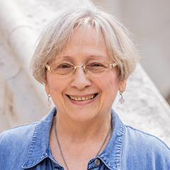 Natural Pharmaceuticals Hepactum FORTE Mosolygó idős nő, rövid ősz hajjal, aranykeretes szemüvegben, világoskék ingben, világos háttér előtt.